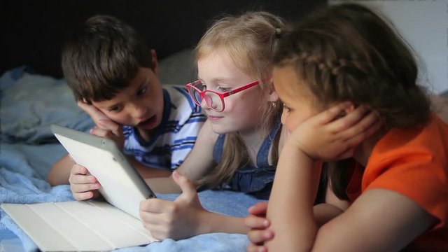 Children are watching a movie on a tablet while lying on a bed