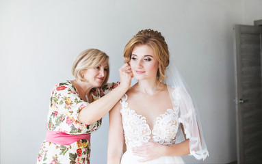 Morning of the bride. Mom helps put on the earrings of a beautiful bride. Mom corrects the wedding hairstyle of the bride. Bride in a beautiful wedding dress