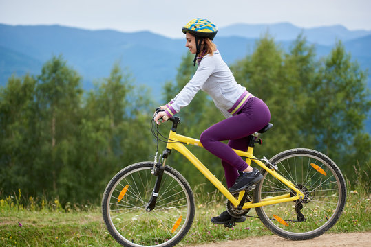 Happy female riding on yellow bicycle on a rural trail in the mountains on summer evening, wearing helmet. Mountains, forests on the blurred background. Outdoor sport activity, lifestyle concept