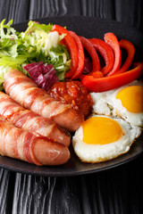 Delicious breakfast of fried sausages wrapped in bacon, eggs and fresh salad close-up on a plate on a table. vertical