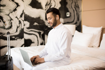 Young busy man with earphones and laptop sitting on bed and typing in hotel room or at home