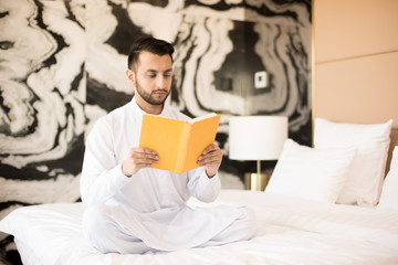 Serious young man with crossed lrgs sitting on white bed and concentrating on reading literature