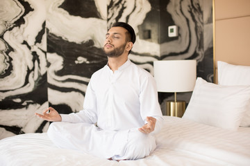 Young man in white sitting on bed in pose of lotus, concentrating and meditating before work