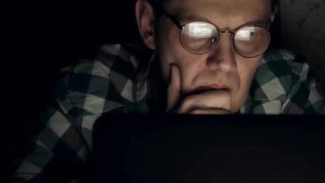 A man in glasses works late at night. He looks worried, he sees on the computer screen in front of him, a close-up