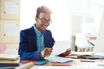 Happy African-american guy in eyeglasses and smart casual reading paper with checked essay after lesson - obrazy, fototapety, plakaty