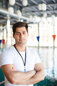 Young swimming instructor with crossed arms looking at camera with pool on background
