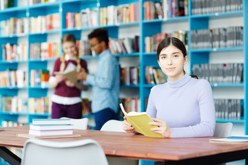 Pretty young woman with open book sitting by desk in library while reading on background of other students