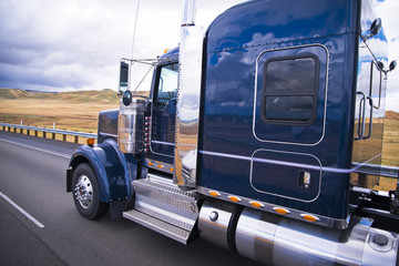Dark blue shiny classic big rig semi truck running on the road with field on background