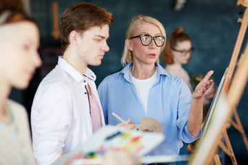 Aged teacher of painting giving advice to guy while commenting his picture on easel at lesson