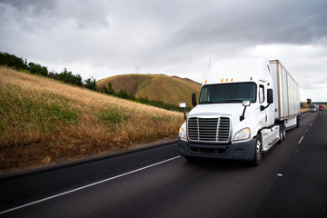 White big rig semi truck with dry van semi trailer driving in straight highway with hill roadside...