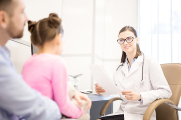 Obraz premium Young smiling clinician with paper talking to father of her little patient during medical appointment