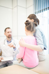 Fototapeta premium Rear view of young woman embracing her daughter during appointment and talk with doctor in clinics