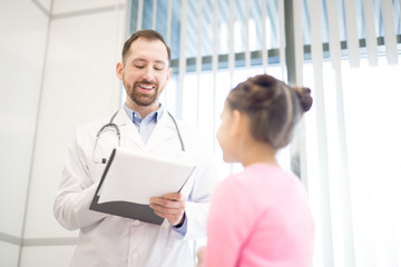 Smiling doctor in whitecoat making notes on paper and giving medical advice to little patient