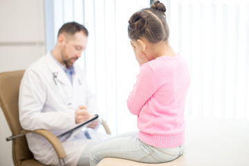 Fototapeta premium Little girl is afraid of bad diagnosis while pediatrician making prescriptions in her medical card