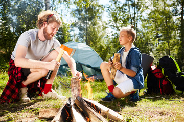 Boy scout helping his father with preparing firewood for campfire on summer weekend