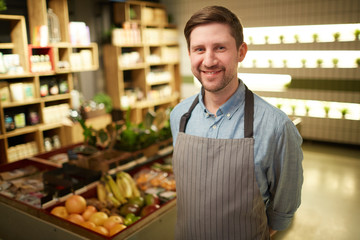Cheerful supermarket assistant in apron looking at you on background of goods