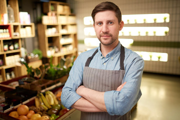 Young man in apron crossing his arms on chest while looking at camera during working day in supermarket