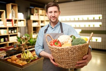 Fototapeta premium Happy supermarket assistant holding big basket with fresh vegetables during work