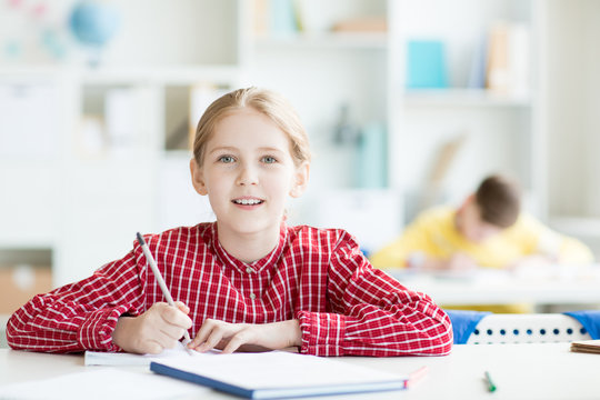 Happy girl looking at camera while carrying out individual task at lesson in classroom