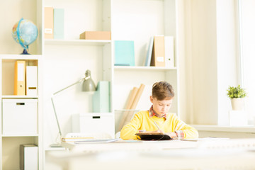 Fototapeta na wymiar Cute young boy making notes or wiriting down home assignment in notebook while sitting by desk