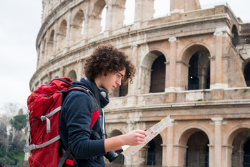 Handsome young traveler looking at tourist map in Rome in front of Colosseum. Backpacker with camera and tourist map