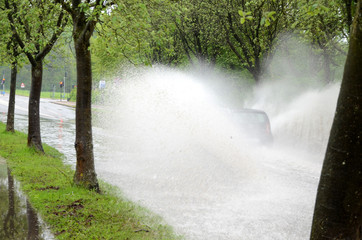 A car hidden in a large splash drives on a road that i flooded because of severe rainfall and bad kept drains.