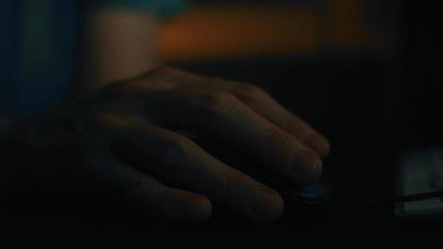 Close up of hand using computer mouse in dark home office interior, low key footage underexposed to emphasize tiredness of man working overtime