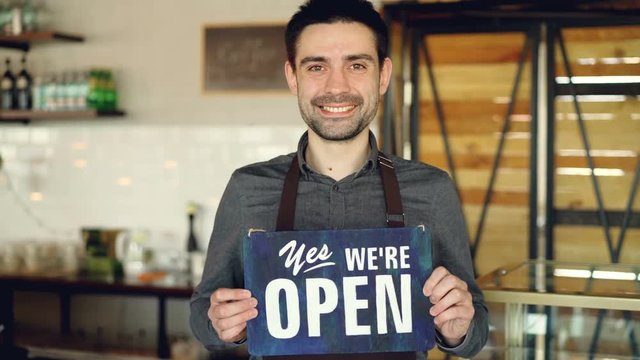 Cheerful handsome man small coffee-house owner is holding "we are open" sign while standing inside coffee shop. Opening new business and people concept.