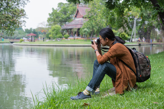male travelers sitting on green grass near pond in public park, he holding camera on hand and take a photo. concept of travel, trip