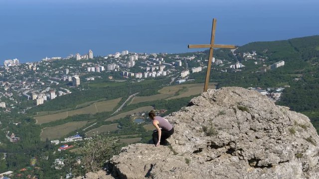 Traveler Woman Sits on Top of a Rocky Mountain. Landscape with mountains and cross on peak on a small town and the sea. Mount Stavrikai. Yalta, Crimea. Achieving the goal. Forward movement, motivation