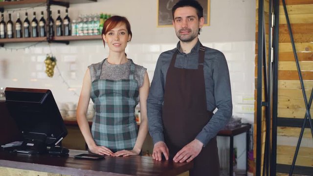 Portrait of two confident waiters in aprons standing at cashier's desk in coffee-house and smiling. Successful business, happy people and food service concept.