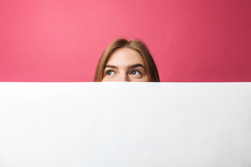 Beautiful young girl peeking out from behind white paper wall, on rose background