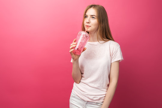 Portrait of beautiful young woman drinking fizzy drink from a bottle with a straw and looking at camera isolated on a pink background, is the text