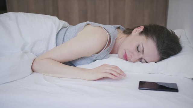 Young beautiful female waking up in bed and looking at smartphone.
