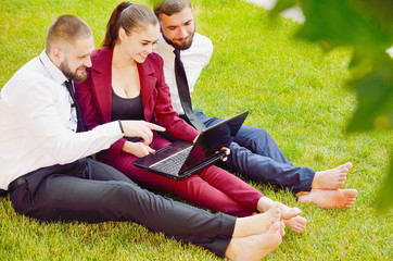 Young office workers with bare feet sit on a green lawn with a laptop and talk. Life style. Teamwork. Guys and the girl. 3.