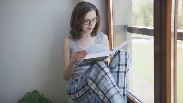 Young beautiful female in eyeglasses and pajamas sitting on window sill at home and reading book.