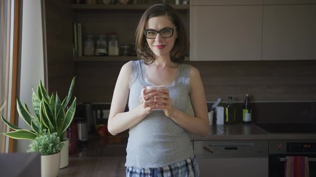 Attractive young female in eyeglasses standing in kitchen holding mug and looking at camera.