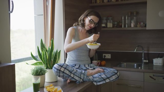 Young beautiful female in pajamas and eyeglasses sitting on top of table in kitchen eating breakfast and smiling.