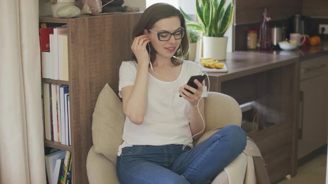 Attractive female in eyeglasses sitting in comfortable armchair at home and using smartphone putting earphones in.