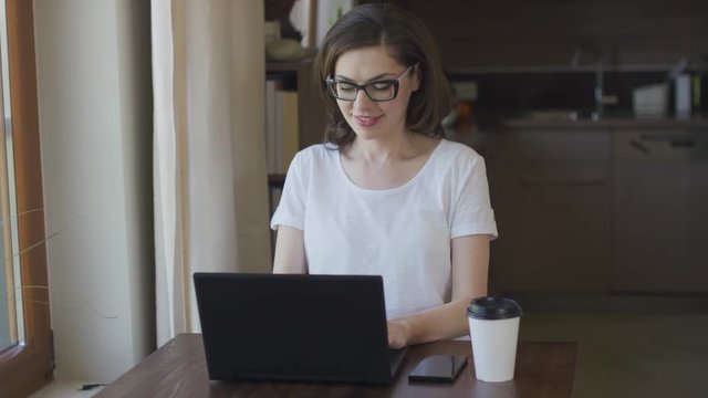 Young beautiful female in eyeglasses sitting at home using laptop and smiling.