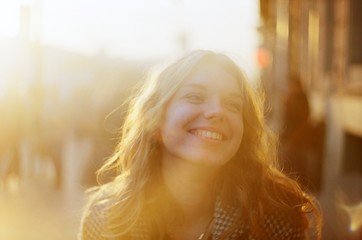 Blonde woman with a beautiful beaming smile backlit by the warm glow of the sun shining down a busy...