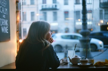Fototapeta Lonely pensive blonde woman thinking in a coffee shop looking through the window at city street. evening obraz
