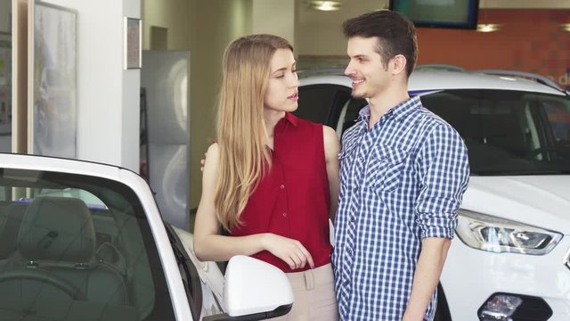 Attractive young happy couple hugging at the dealership salon while examining cars for sale. Handsome young man and his beautiful girlfriend choosing a new auto to buy.