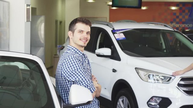 Attractive young man posing near his newly bought automobile showing car keys to the camera. Happy male driver at the dealership showroom. Consumerism, rental, driving concept.