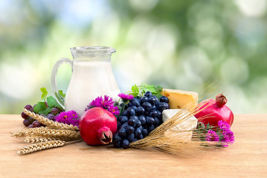 Grapes, garnets, milk in a pitcher, yellow and white cheese, pink flowers, barley and wheat on wooden table on a blur nature background