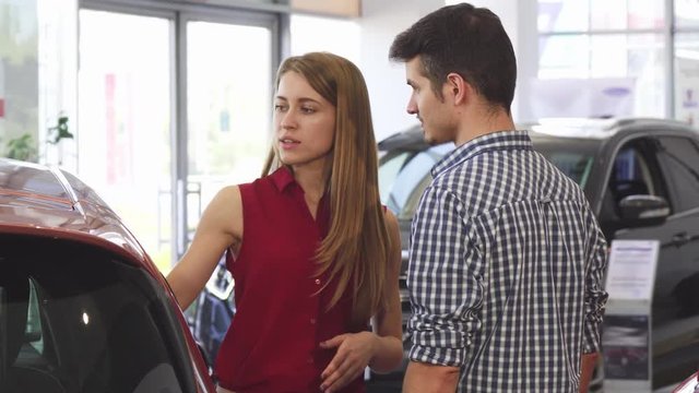 Gorgeous young woman talking to her boyfriend while choosing new car together at the dealership. Happy couple examining cars for sale at the dealership salon. Buying, purchase, sales.