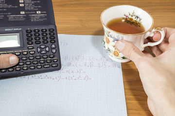 A student is doing math homework with a calculator and an hot cup of tea on a wood table