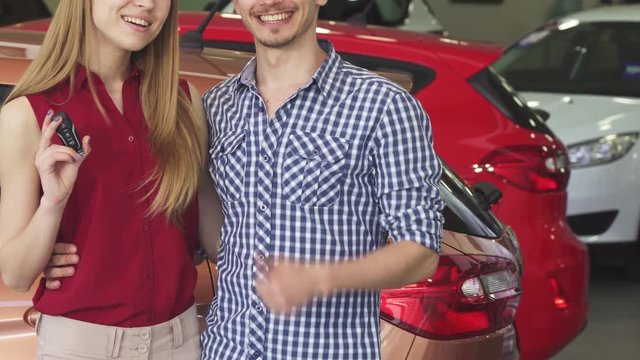 Cropped shot of a happy couple buying a new automobile together at the dealership showroom showing thumbs up and car keys. People buying auto. Vehicle ownership.