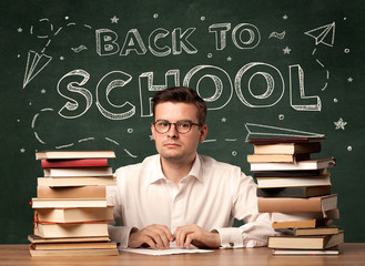 A young teacher in glasses sitting at classroom desk with pile of books in front of blackboard saying back to school drawing concept.