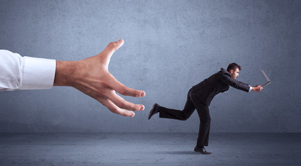 Young miniature businessman running from a big hand with blueish grey background